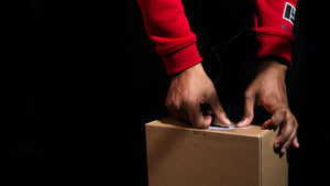 Male hands opening a brown box with red sweatshirt sleeves showing on a black background. Imaged used as new product drop banner message on blueigloo.ca