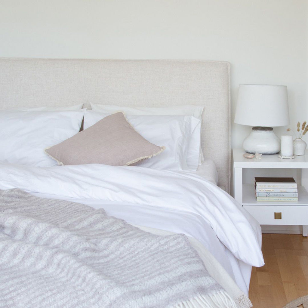 Julia Storage Bed, Cream. Showing bed dressed with a white duvet & varied pillows with a striped throw at the bottom.  There is a  white end table with a white lamp, books, and other decorative objects on top to the right.  Shop storage beds and furniture online at blueigloo.ca