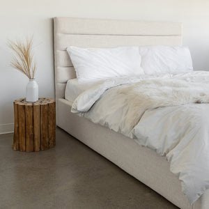 Justin Storage Bed-Tall, Cream by Style In Form. Lifestyle shot of bed in white room with concrete floor, bed is dressed with duvet, pillows and fur throw, there is a wood stump end table on the left side with a white vase on it. View from left end side of the bed. Shop vintage & new home décor, lighting & home furnishings as well as novelty gifts & pet accessories online at blueigloo.ca